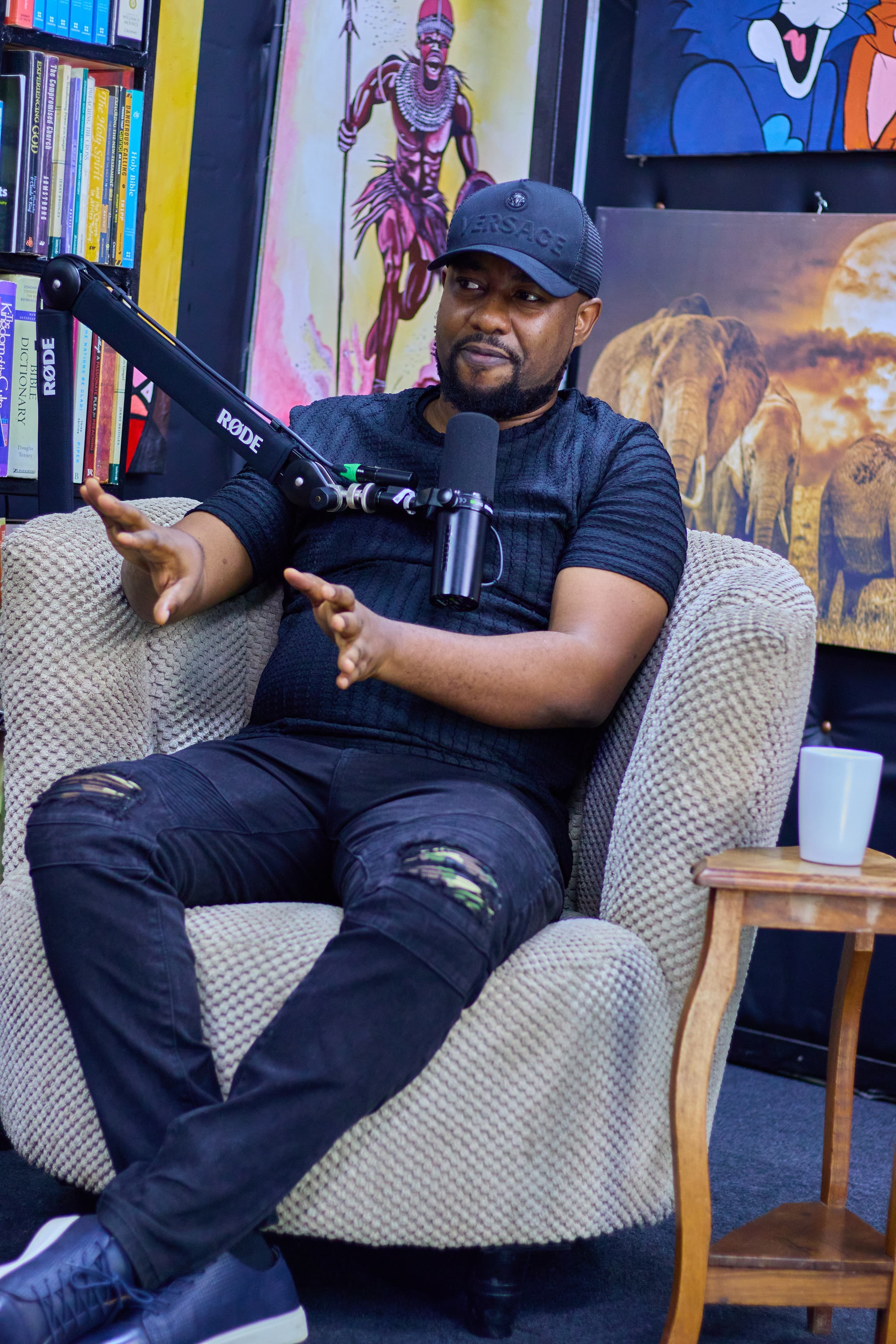Keith Muoki seated in an armchair on a podcast set, gesturing with both hands as he speaks into a studio microphone