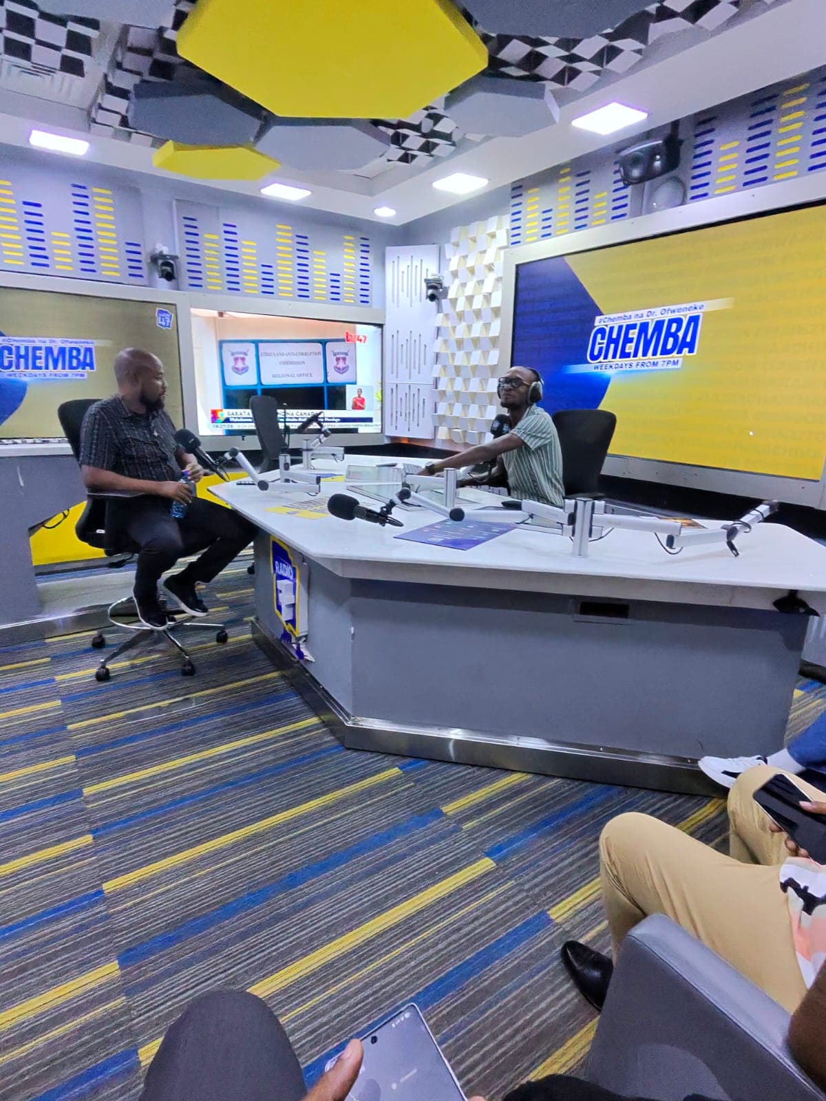 Keith Muoki seated across the desk from a presenter in the Radio 47 studio, with the #Chemba na Dr. Ofweneke branding on the screens behind them