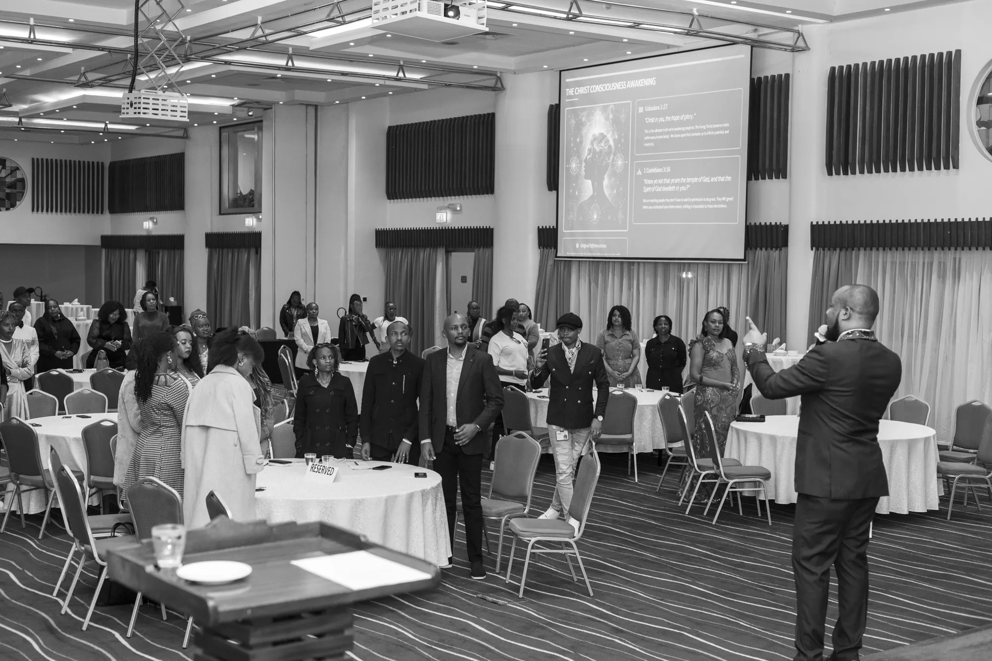 Black-and-white photograph of Keith Muoki, microphone in hand, addressing a full room of standing delegates at banquet tables, with a presentation slide projected behind them